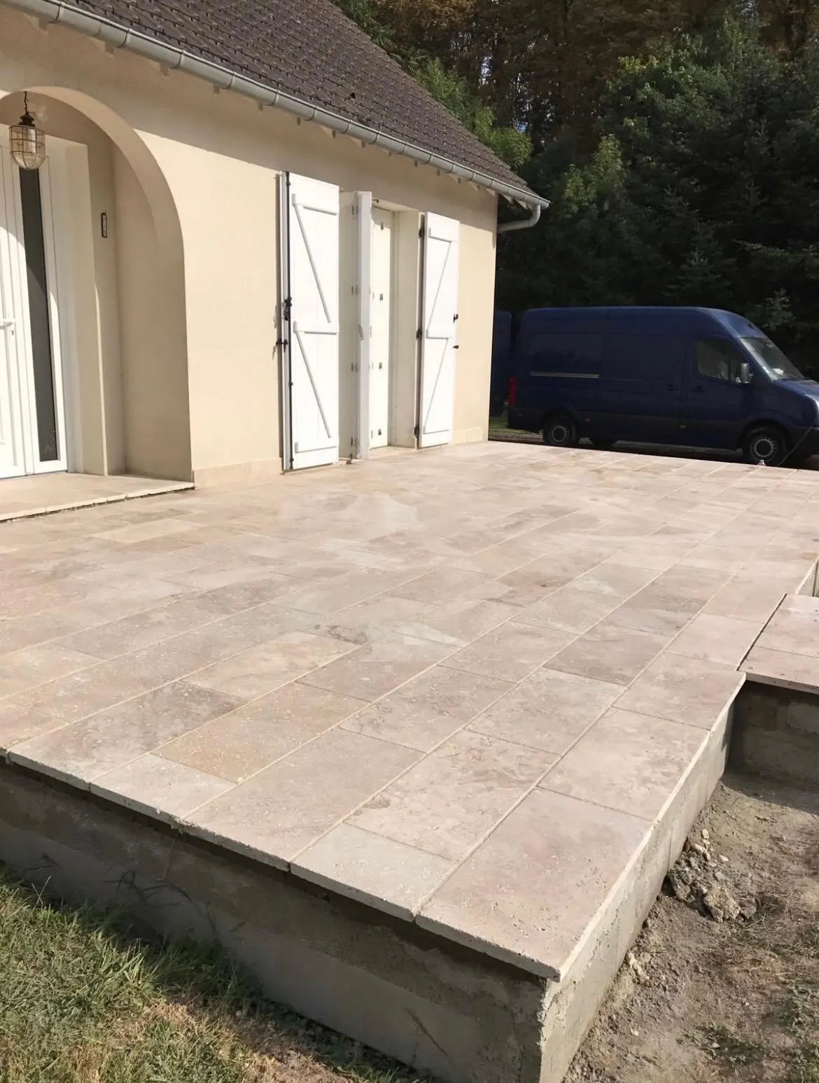 Terrasse surélevée en dalles de travertin posées devant une maison aux volets blancs