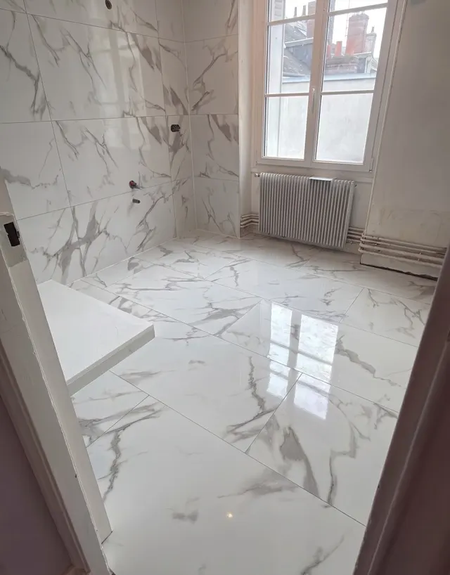 Salle de bains en grands carreaux effet marbre blanc veiné, au sol et aux murs, devant une fenêtre