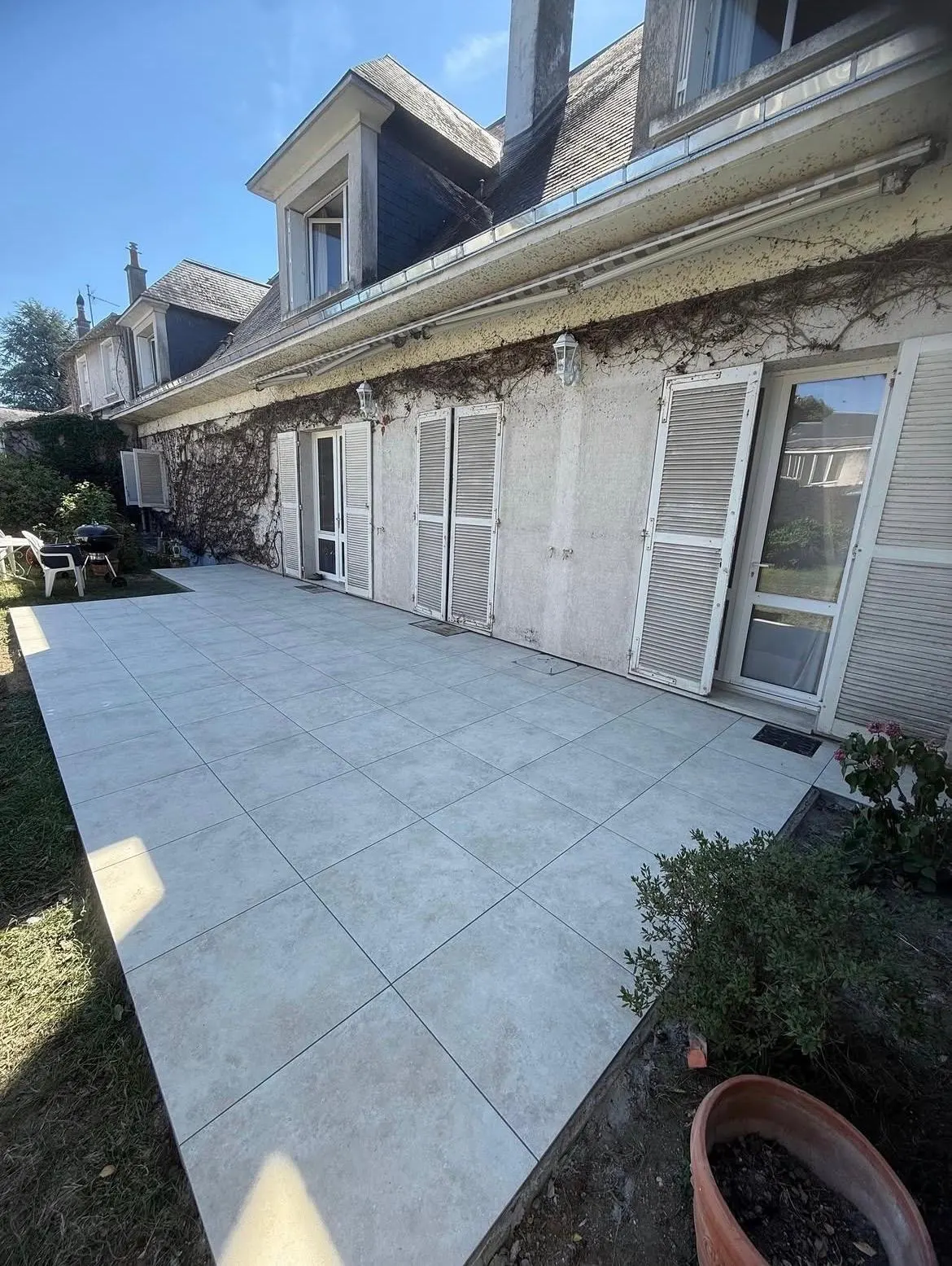 Grande terrasse en dalles claires grand format le long d'une maison aux volets blancs