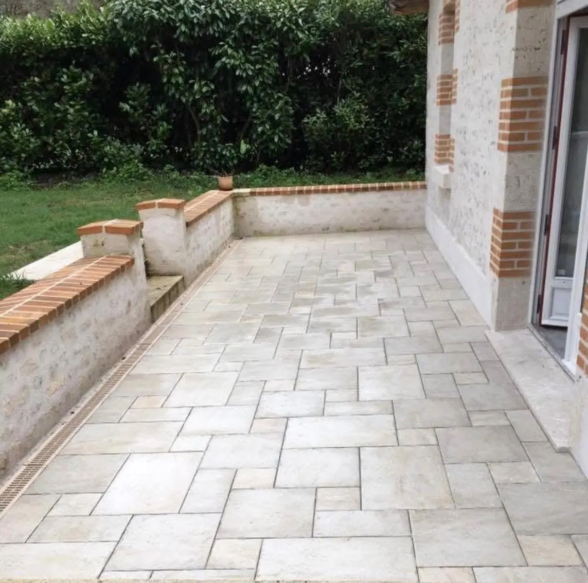 Terrasse en dalles de travertin bordée de murets en pierre et briques, devant une haie